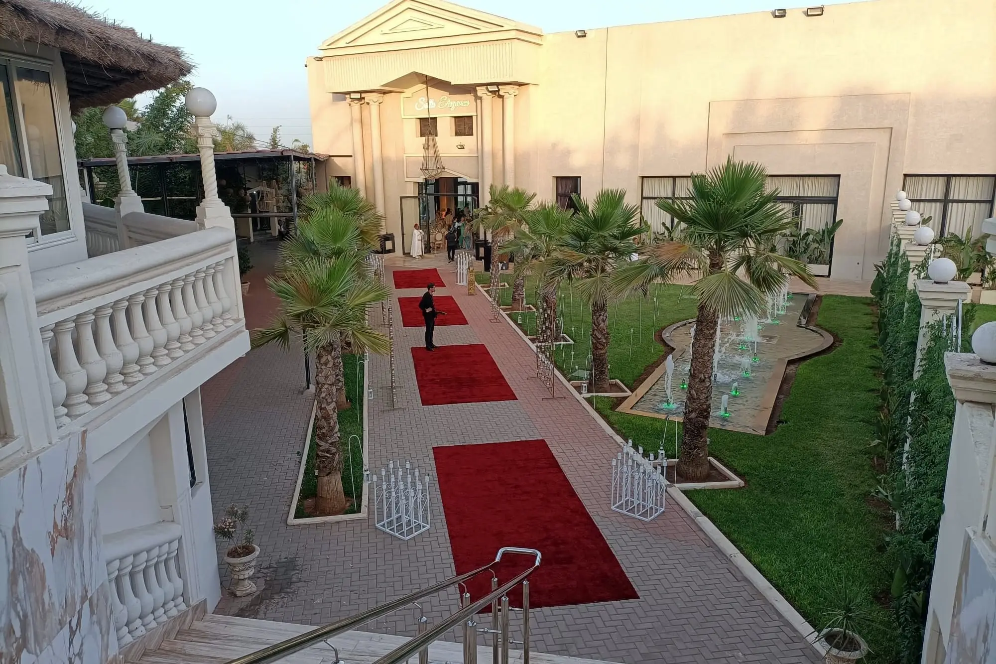 Allée bordée de palmiers et de balustrades blanches, tapis rouges posés vers le pavillon « Salle Élégance »