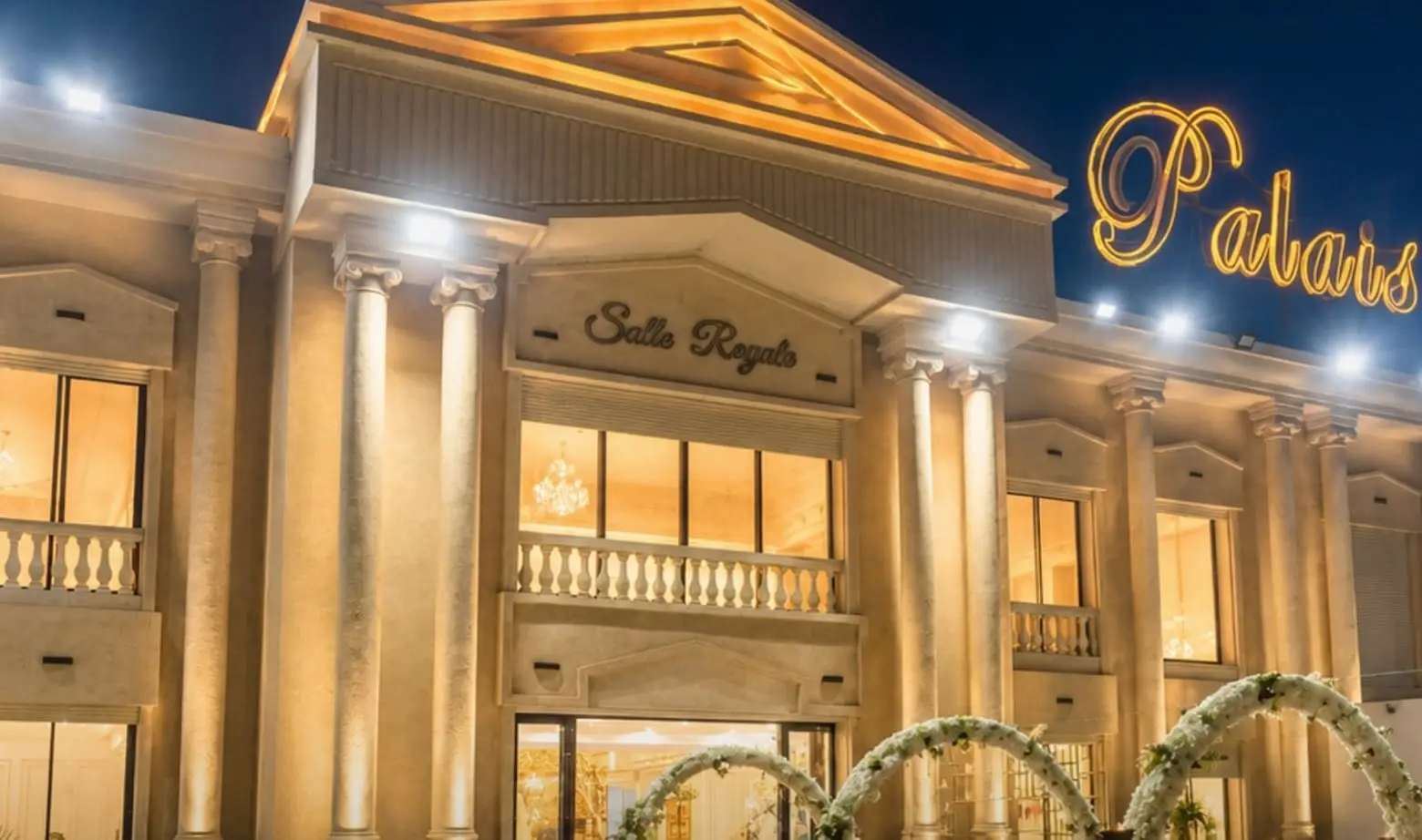 The façade at blue hour: the “Salle Royale” pediment, the lit “Palais Elga” sign, floral arches