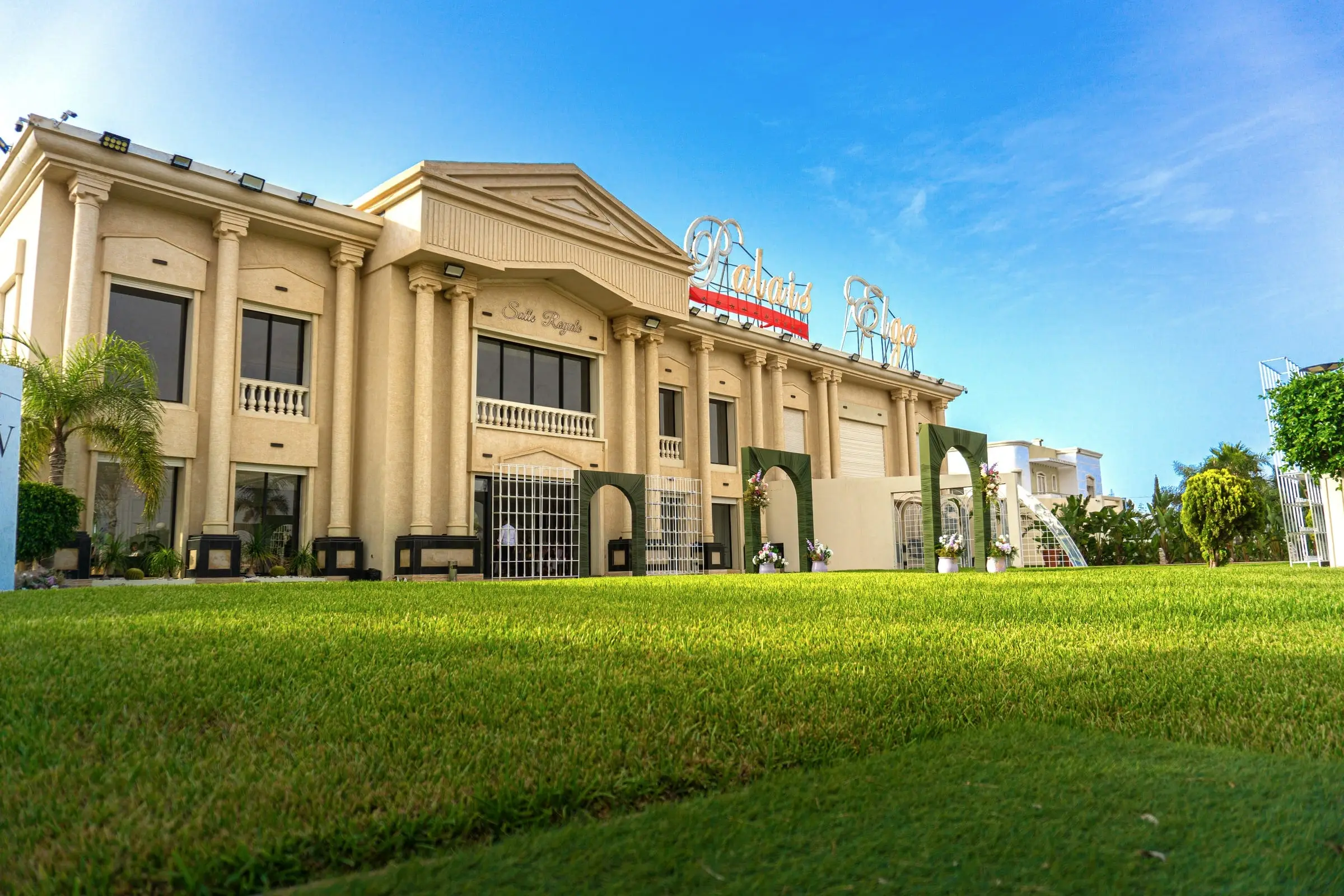 The neoclassical façade of Palais Elga by day: columns, pediment and rooftop sign above the lawn