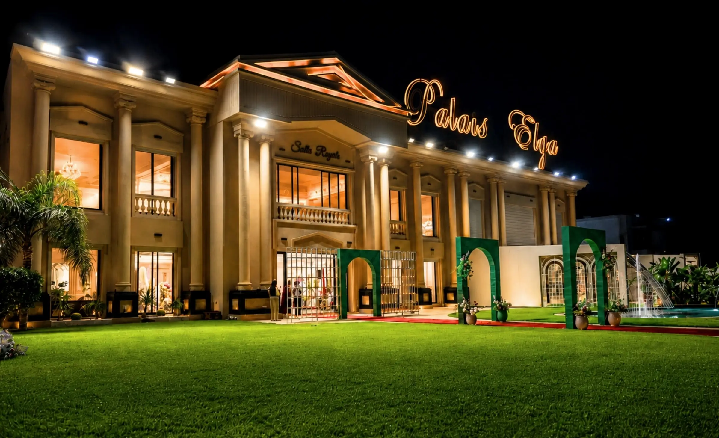 The same façade lit at night, the “Palais Elga” sign glowing on the roof