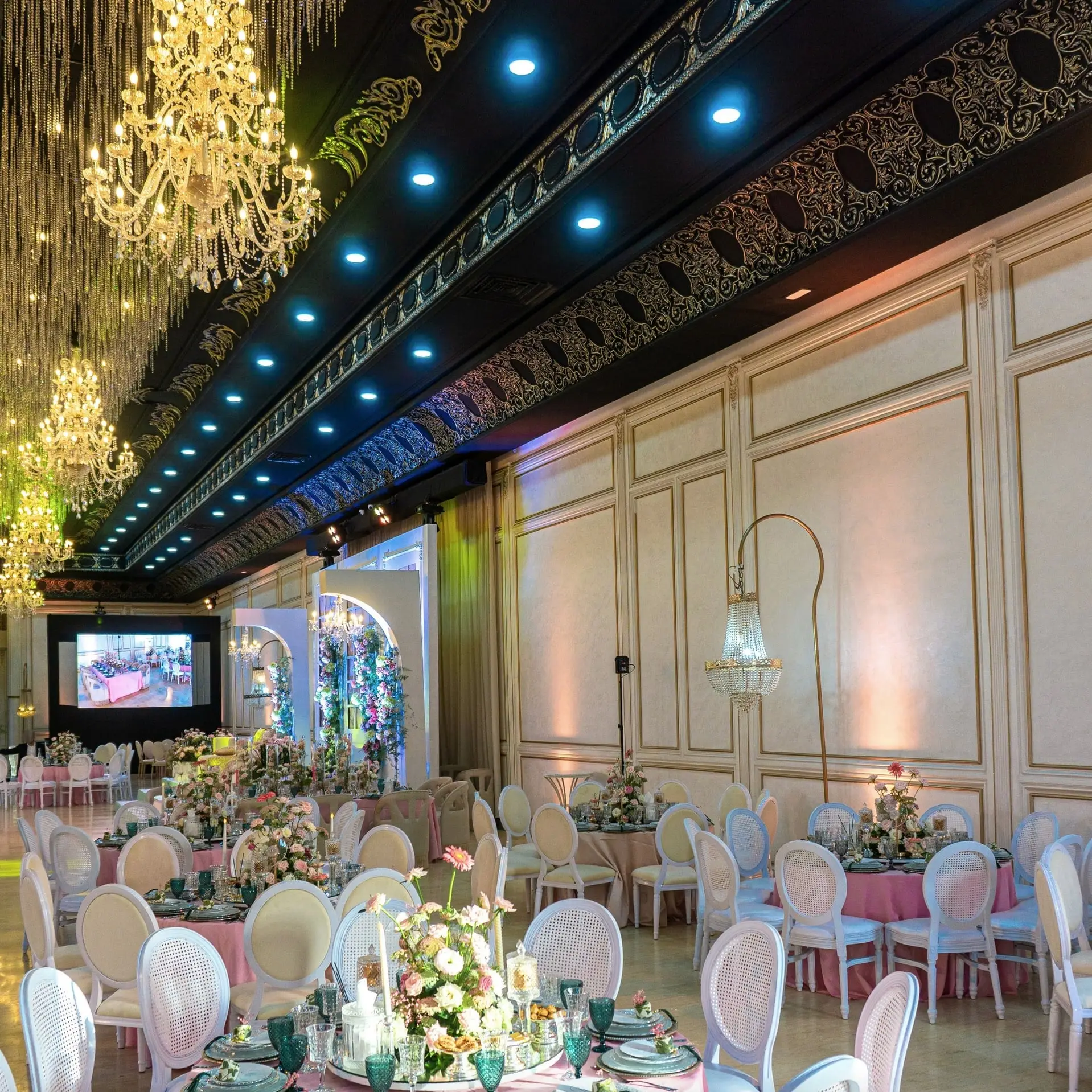 Black-ceilinged hall laid for dinner: pink cloths, white chairs, floral centrepieces, chandeliers