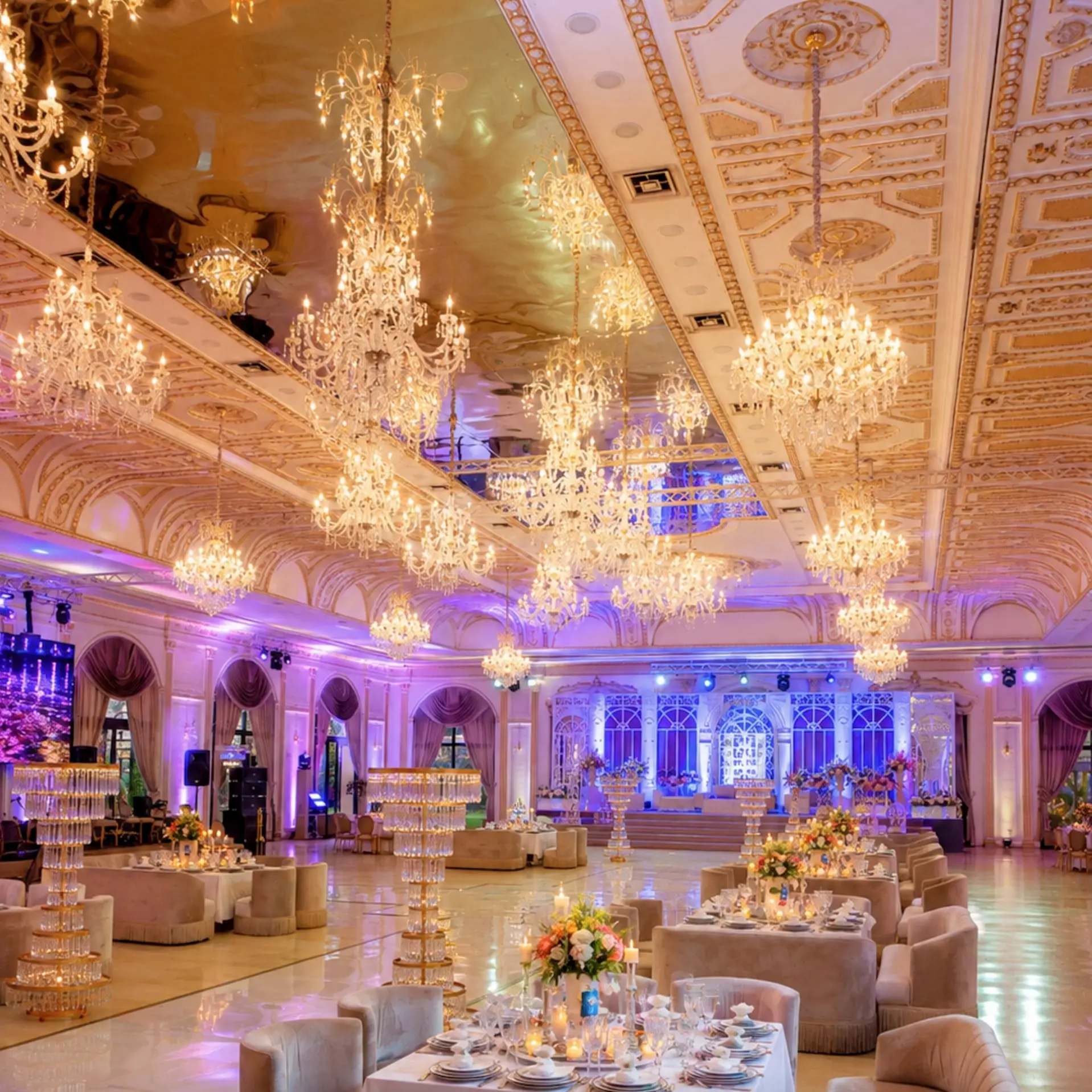 White-and-gold hall with dozens of chandeliers, lounge tables, violet light toward the stage